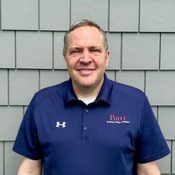 Middle-aged man with gray hair wearing a dark blue polo shirt with Under Armour and Dana logos, photographed outdoors against a gray shingle wall