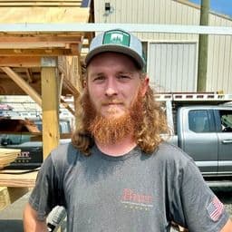 Man with long red beard wearing green cap and gray t-shirt standing in front of wooden structure and vehicles