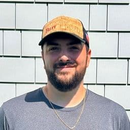 Man wearing tan baseball cap and gray t-shirt smiling at camera against white brick wall background